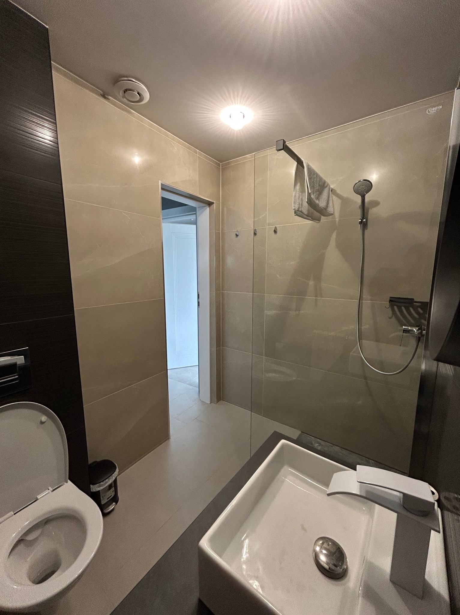 Nowoczesna łazienka po remoncie: beżowe płytki, kabina prysznicowa ze szklaną ścianą, biała umywalka i toaleta. Widok na otwarte drzwi.