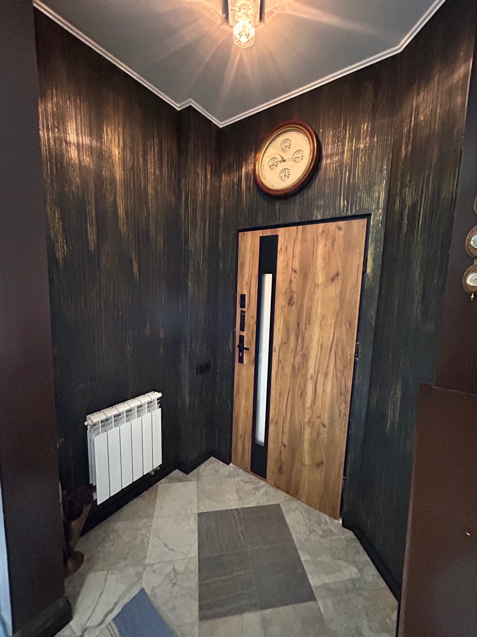 Elegancki przedpokój z dekoracyjną ścianą w odcieniach czerni i złota, drewnianymi drzwiami z czarnymi akcentami i marmurową podłogą.