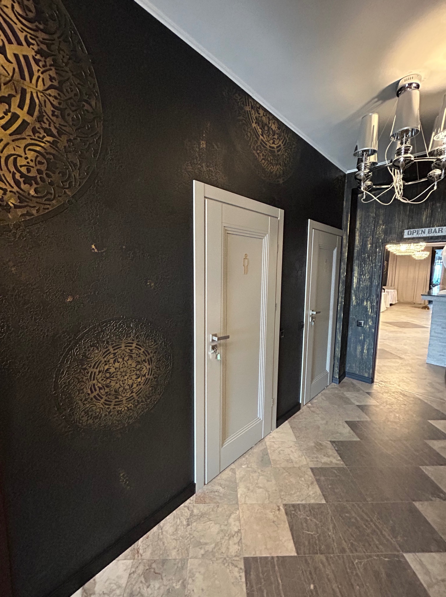 Eleganckie wykończenie wnętrza: ciemna ściana z ornamentami, jasne drzwi z oznaczeniem toalety, wzorzysta podłoga i widok na salę z żyrandolem.
