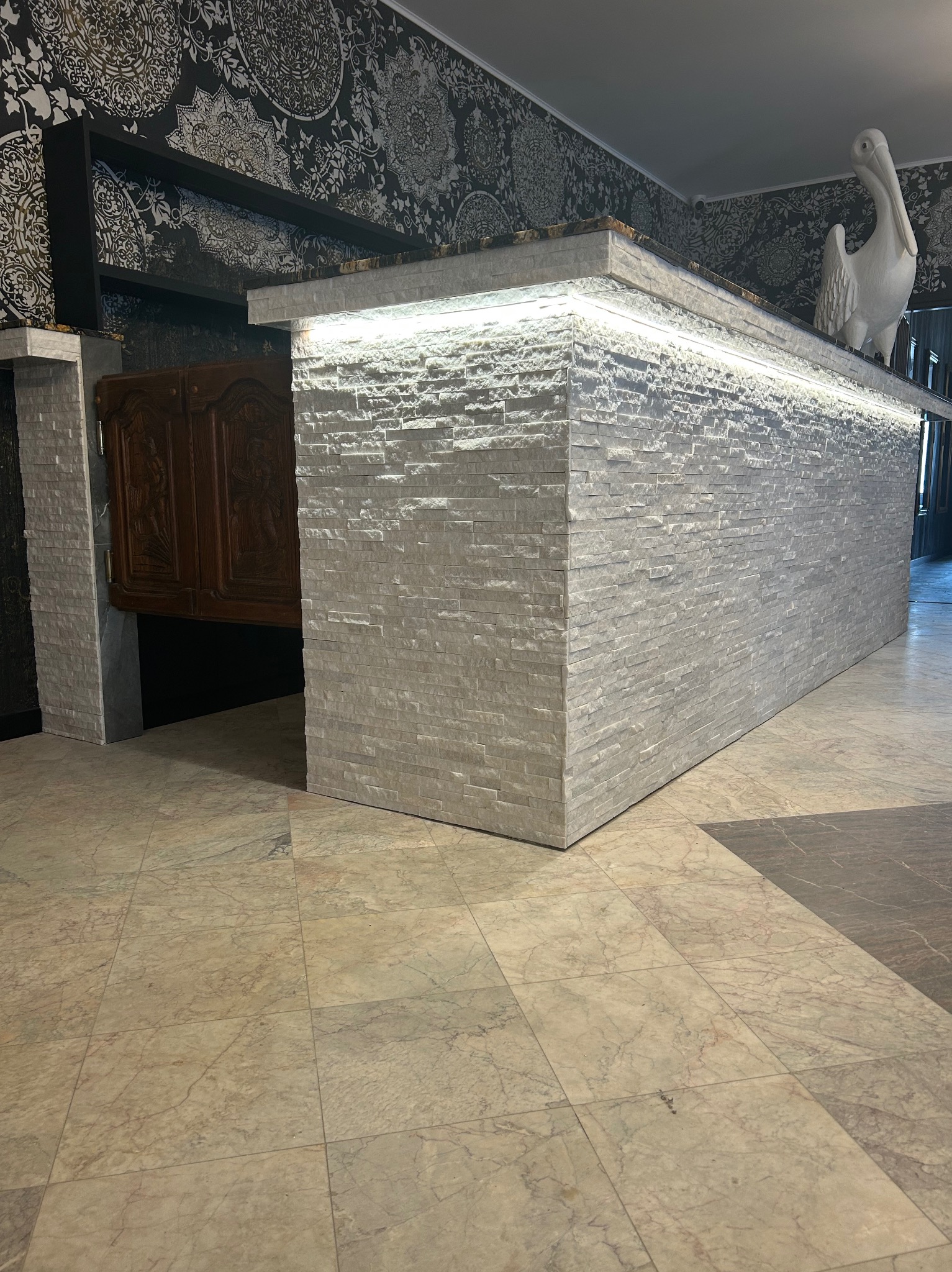 Wykończona wyspa kuchenna z białej cegły z oświetleniem LED i dekoracyjnym pelikanem, wzorzysta tapeta w tle. Marmurowe płytki podłogowe.