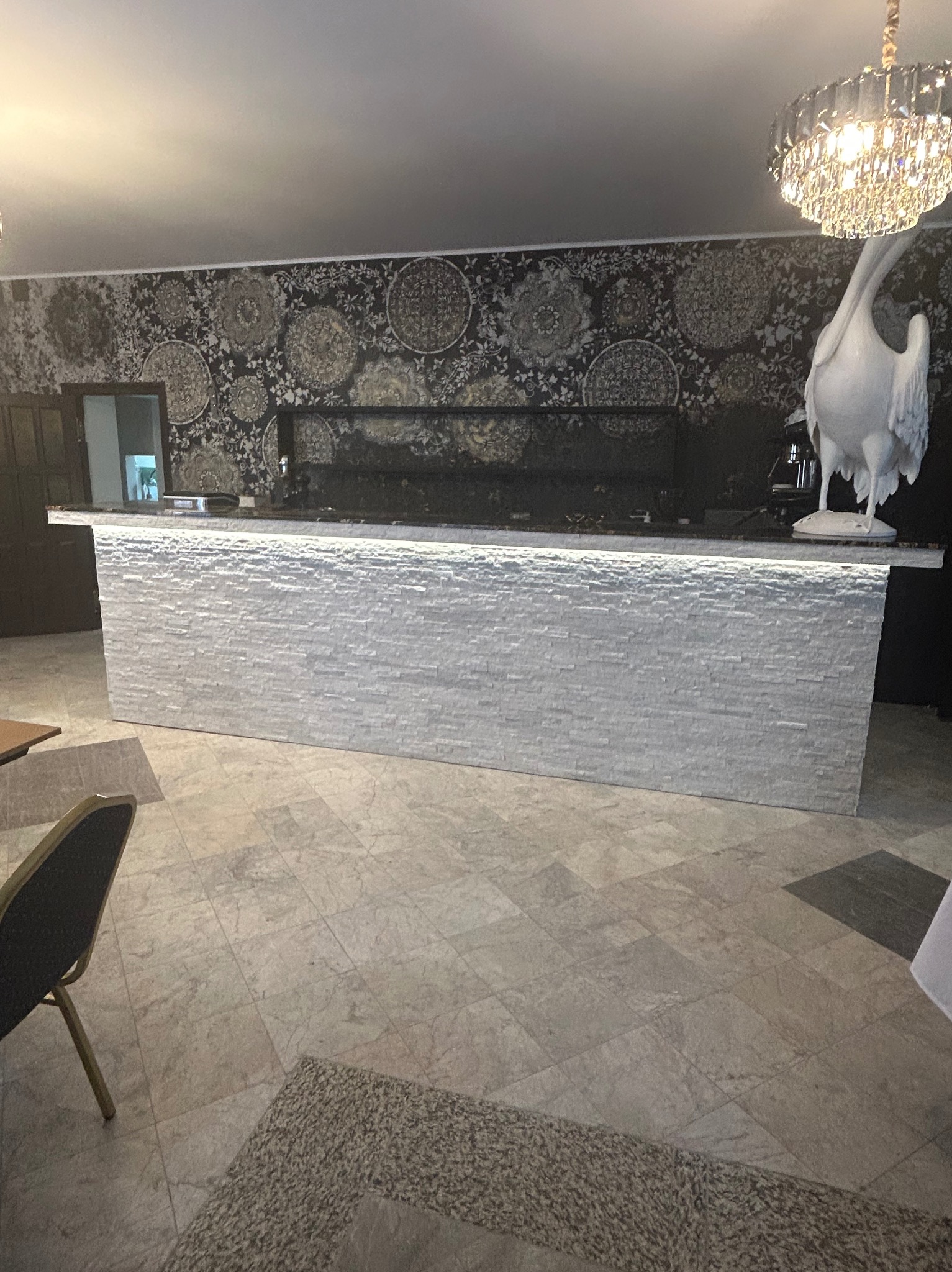 Biały bar z podświetleniem i dekoracyjnym kamieniem na tle wzorzystej tapety i kryształowego żyrandola. Wykończenie wnętrza w eleganckim stylu.