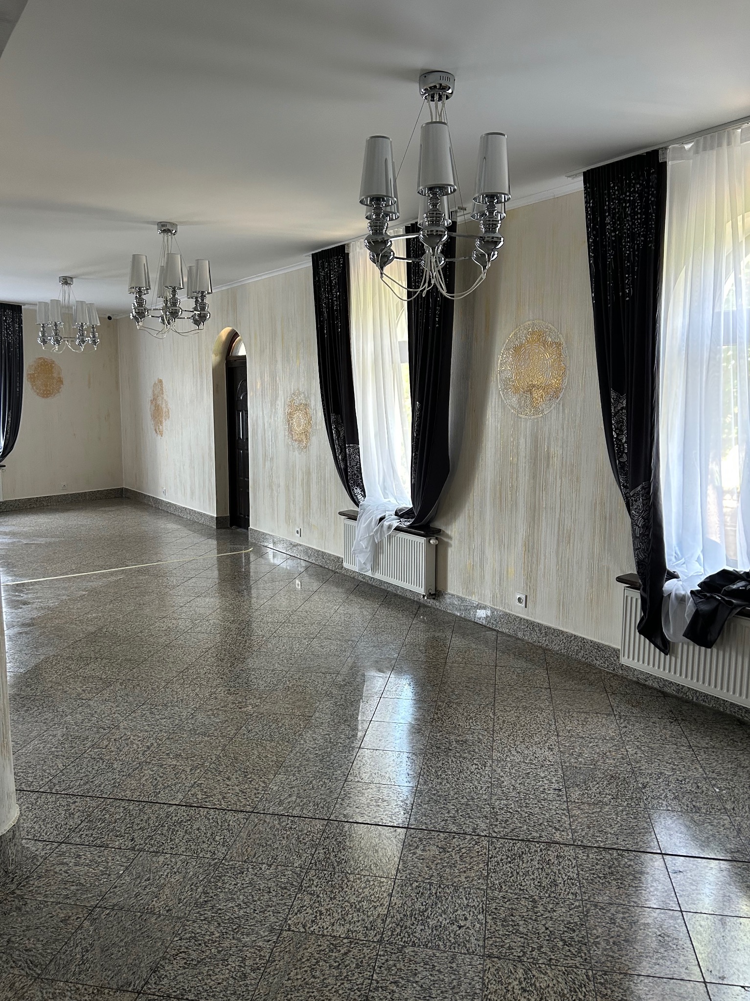 Eleganckie wnętrze sali balowej z błyszczącą, granitową posadzką, dekoracyjnymi lampami i ścianami z subtelnymi zdobieniami. Czarne i białe zasłony dodają szyku.