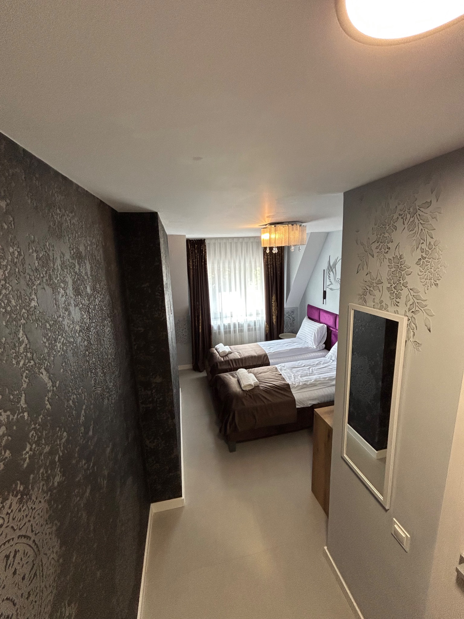 Pokój hotelowy z dwoma łóżkami, ciemną, teksturowaną ścianą i dekoracyjnym wzorem kwiatowym na jasnej ścianie. Widoczne lustro i oświetlenie sufitowe.