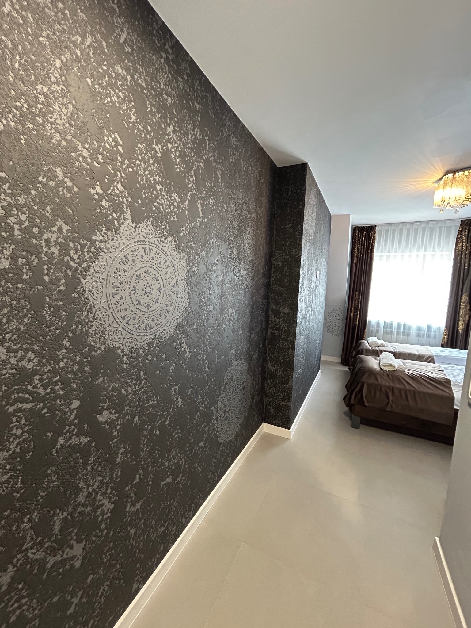 Stylowa ściana z dekoracyjnym wzorem w odcieniach szarości w sypialni. Widoczne dwa łóżka z brązową pościelą i białe ręczniki. Całość dopełnia jasna podłoga.