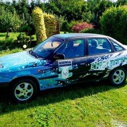 Epicdot - Oryginalnie pomalowany samochód Opel Astra w Koszalinie, z motywami sportowymi i gwiazdami, zaparkowany na trawniku w otoczeniu zieleni.