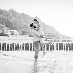 Epicdot - Czarno-białe zdjęcie zakochanej pary na plaży: mężczyzna trzyma kobietę w ramionach na tle falochronu i łodzi na piasku, tworząc romantyczną atmosferę.