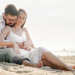 Epicdot - Zakochana para siedzi na piasku plaży w słoneczny dzień. Mężczyzna obejmuje kobietę, oboje ubrani w jasne stroje, z zamyślonymi wyrazami twarzy. W tle widać morze.