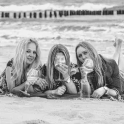 Trzy kobiety leżą na plaży, pijąc wino z kieliszków na tle morza i falochronów. Zdjęcie czarno-białe, z wyraźnym pierwszym planem piasku.