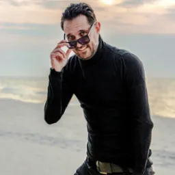 Mężczyzna z brodą, w czarnym golfie i okularach przeciwsłonecznych, uśmiecha się na tle plaży o zachodzie słońca. Okulary lekko zsunięte, odsłaniają oczy.