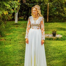 Epicdot - Uśmiechnięta panna młoda w koronkowej sukni z długim rękawem, stojąca na trawie w plenerze. W tle drzewa i dekoracje ogrodowe.