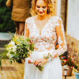 Epicdot - Uśmiechnięta panna młoda w koronkowej sukni trzyma bukiet, w tle mężczyzna i balony. Detal: kwiatowa ozdoba na nadgarstku.