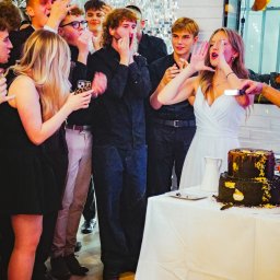 Epicdot - Panna młoda kroi tort weselny, otoczona gośćmi wyrażającymi radość. W tle srebrna ściana i balony. Ujęcie z bliska, dynamiczne.