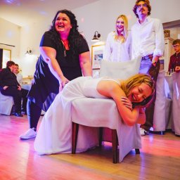 Epicdot - Radosna zabawa weselna: panna młoda leży na krześle, a goście weselni śmieją się w tle, oświetleni kolorowymi światłami.