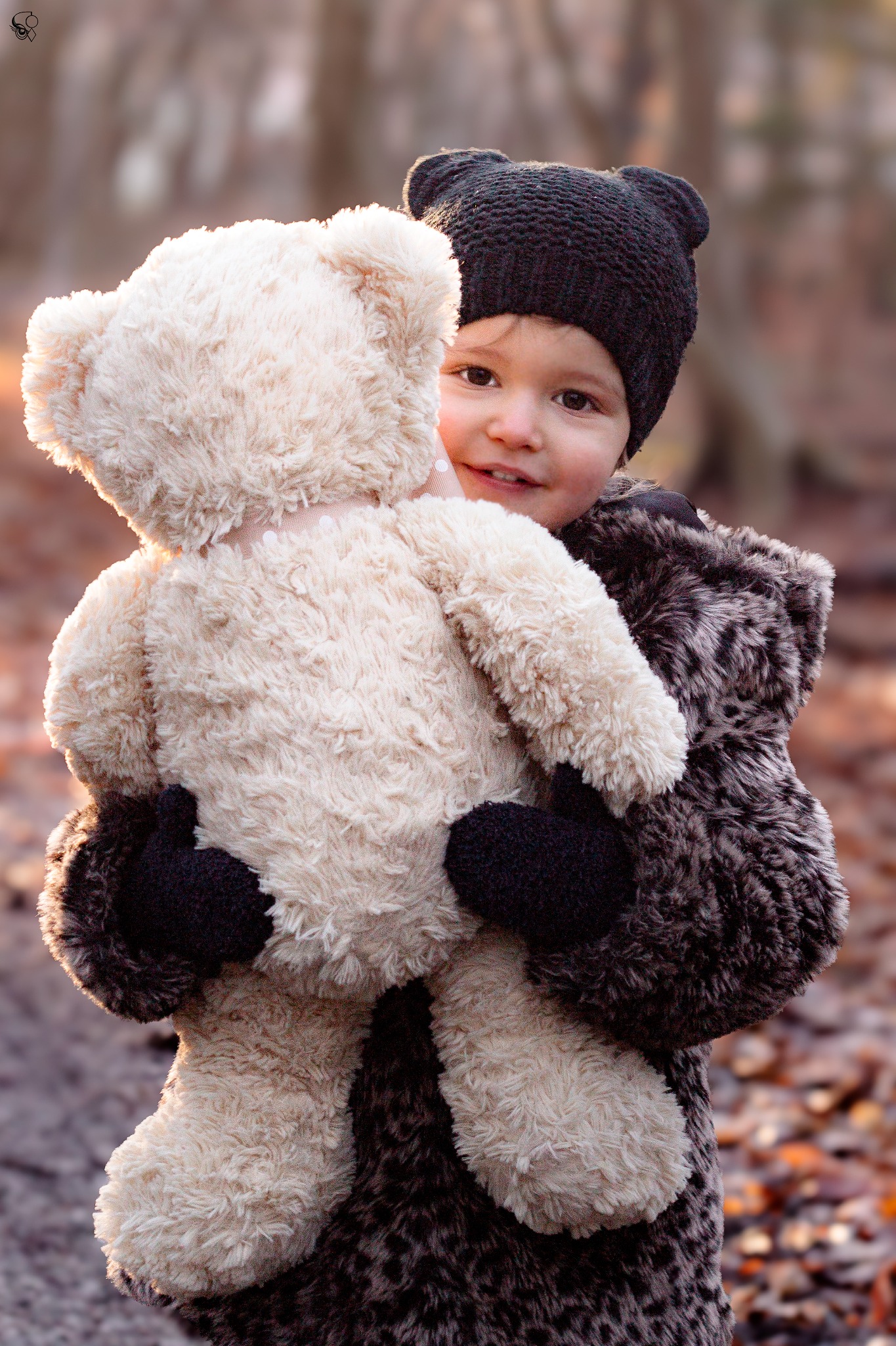 Uśmiechnięta dziewczynka w czapce z uszkami przytula pluszowego misia na tle jesiennego lasu, ciepłe światło.