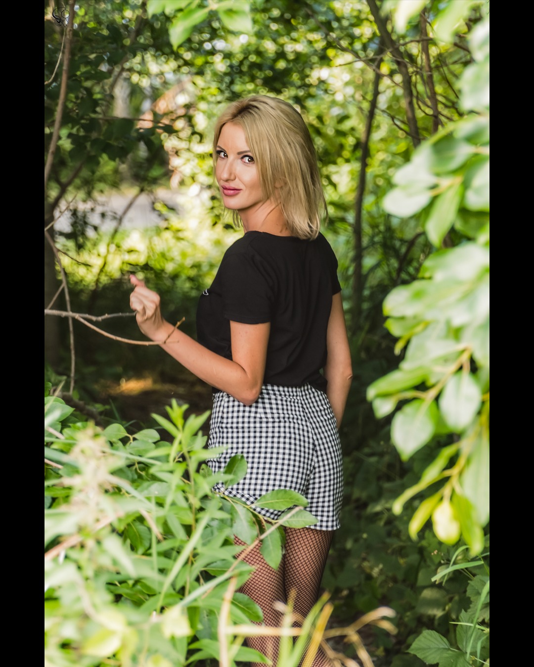 Kobieta z blond włosami, odwrócona przez ramię, w czarnej koszulce i kraciastych szortach, pozuje w zielonym otoczeniu, z delikatnym uśmiechem i spojrzeniem w obiektyw.
