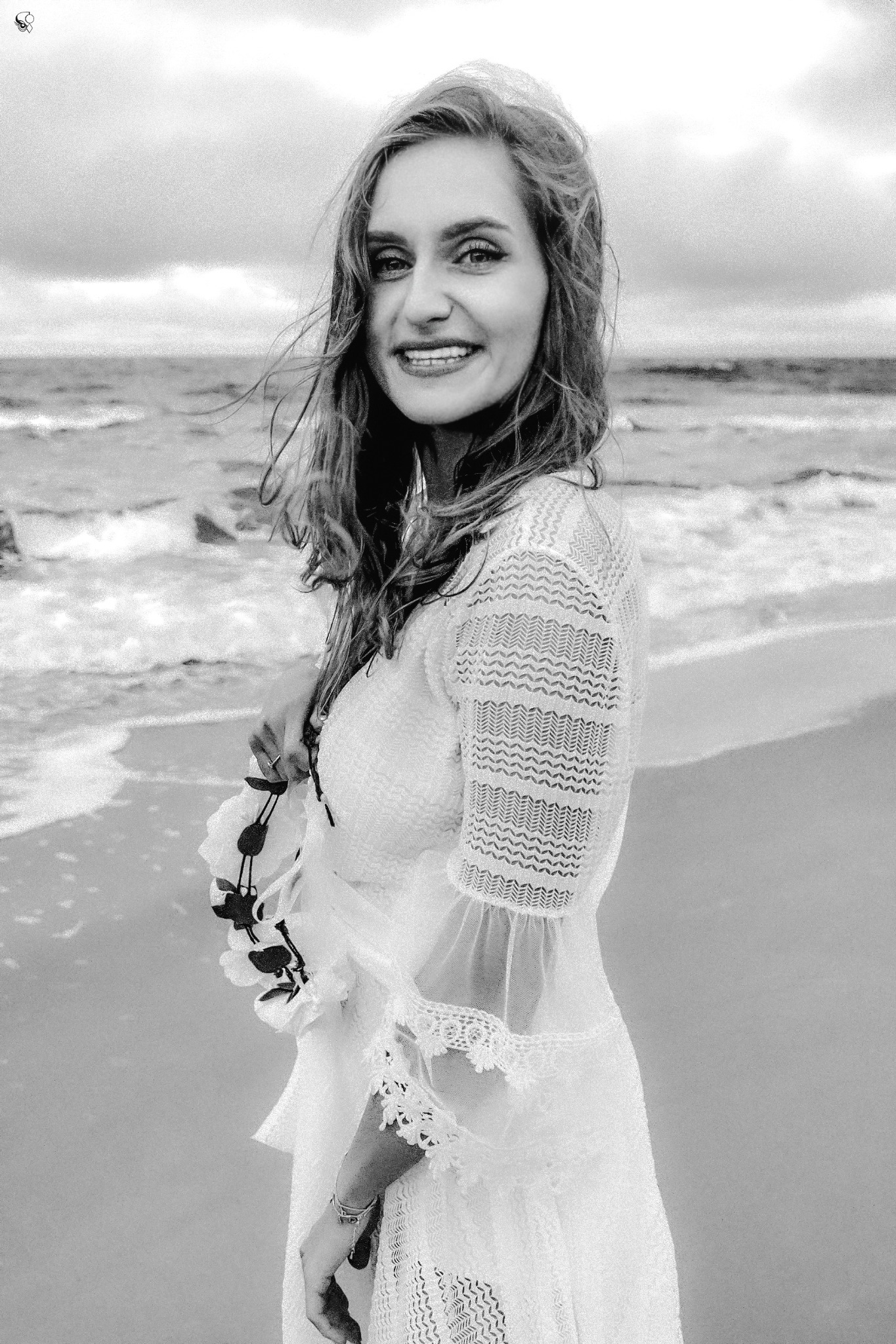 Portret uśmiechniętej kobiety w koronkowej sukience na tle morza. Zdjęcie czarno-białe, stylizowane, z wiankiem w ręku, podkreślające naturalność i subtelność.