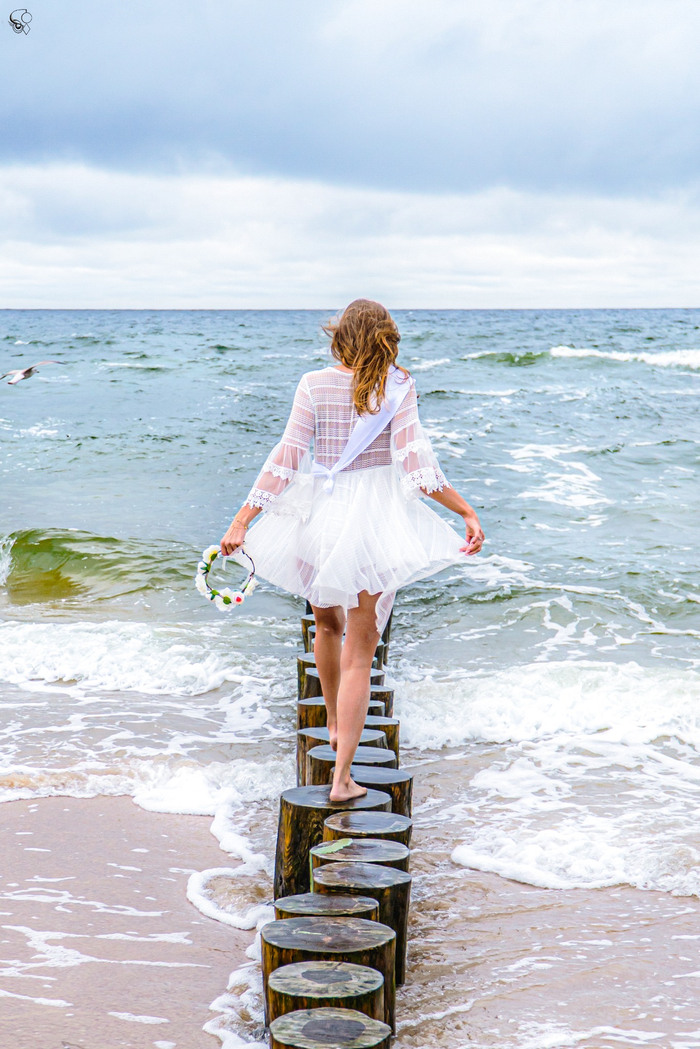 Kobieta w białej sukience i z wiankiem idzie po drewnianych palach w morzu, z widokiem na fale i niebo. W tle przelatuje mewa.