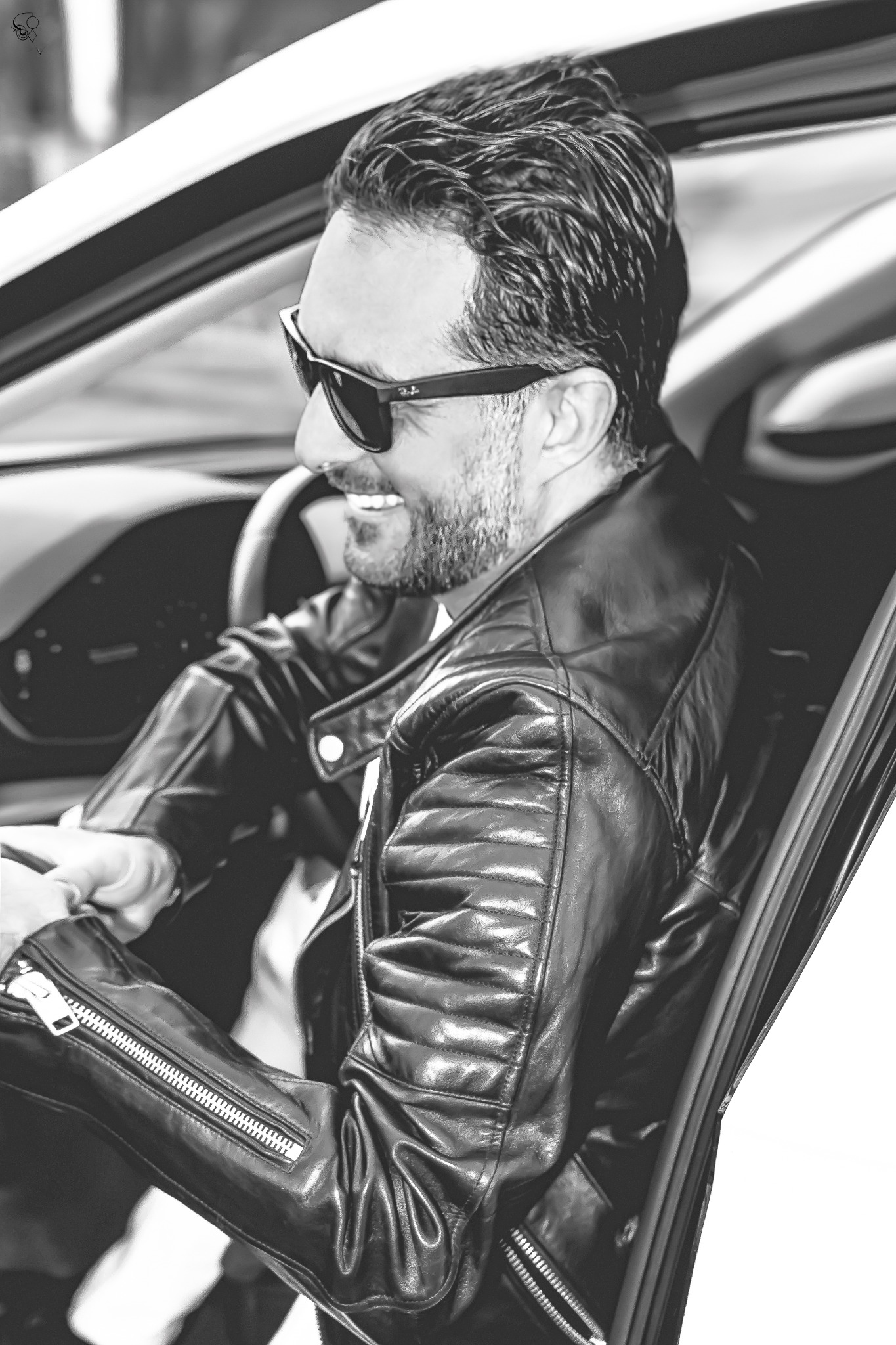 Czarno-białe zdjęcie uśmiechniętego mężczyzny w skórzanej kurtce i okularach przeciwsłonecznych, siedzącego w samochodzie. Widoczna kierownica i fragment deski rozdzielczej.