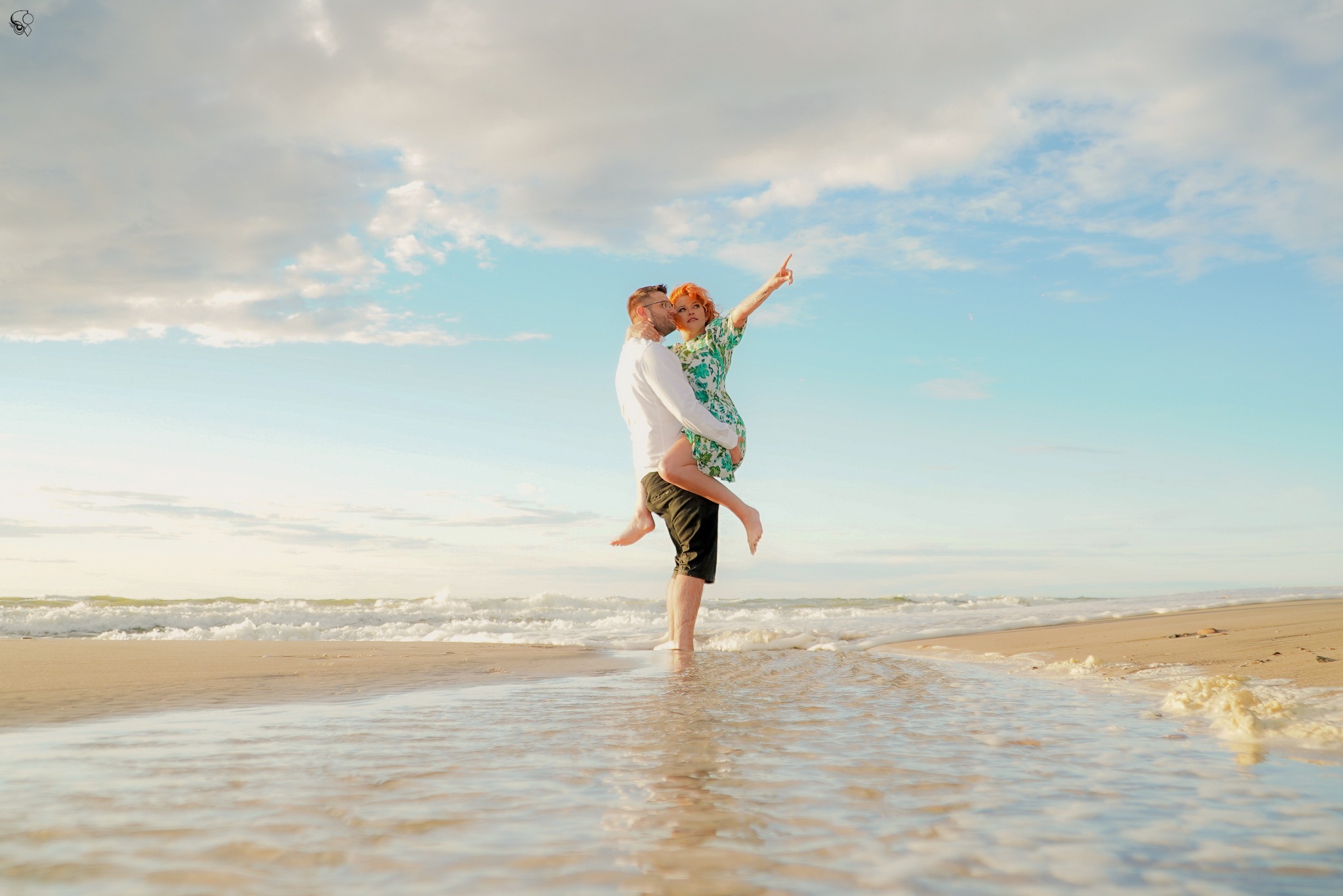 Para na plaży: mężczyzna trzyma kobietę w ramionach, ona wskazuje palcem w górę. Delikatne fale obmywają ich stopy, a w tle rozciąga się błękitne niebo z chmurami.