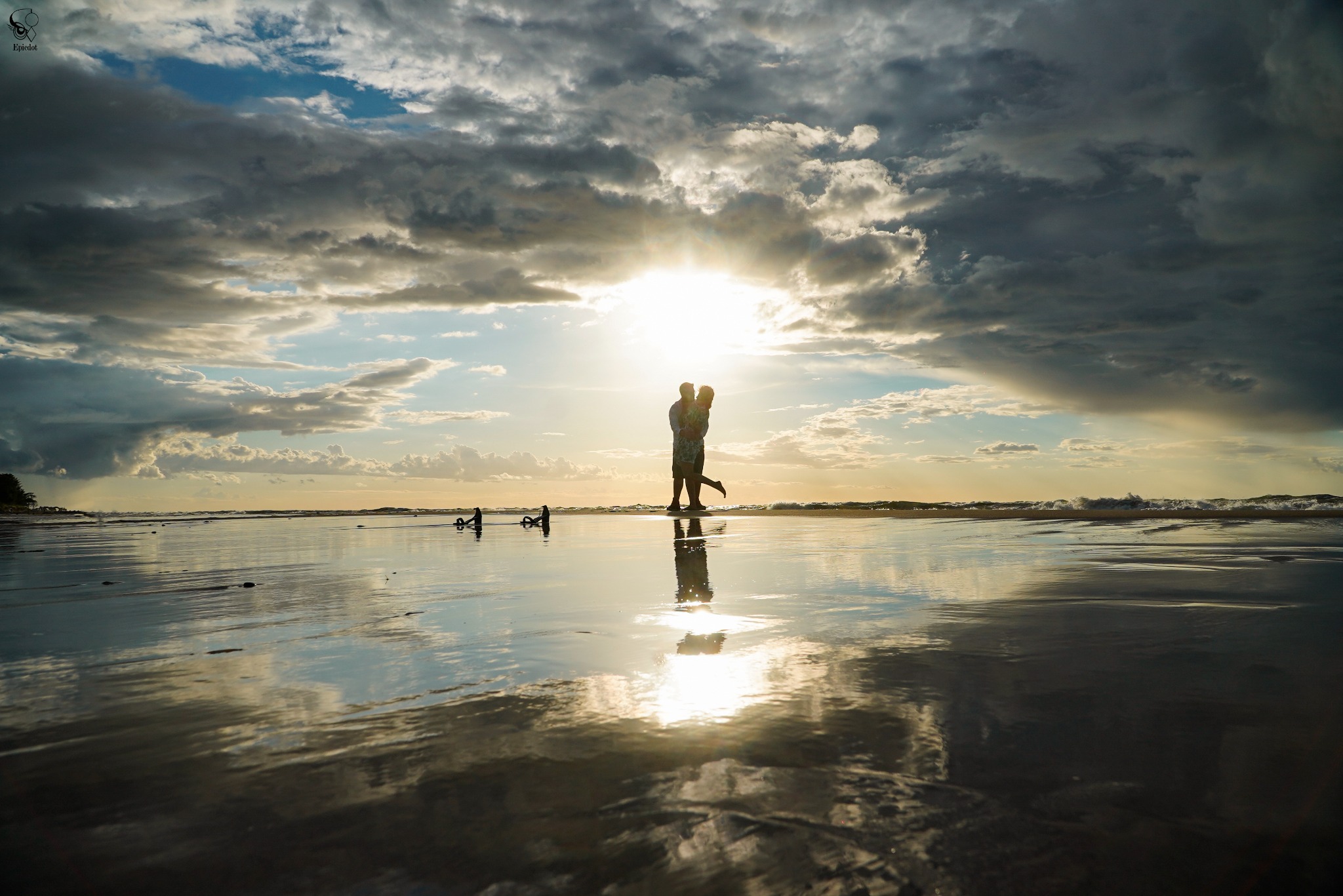 Para w objęciach na plaży o zachodzie słońca, ich sylwetki odbijają się w mokrym piasku. Dramatyczne chmury i niski kąt ujęcia dodają romantyzmu. W tle widać spokojne morze.