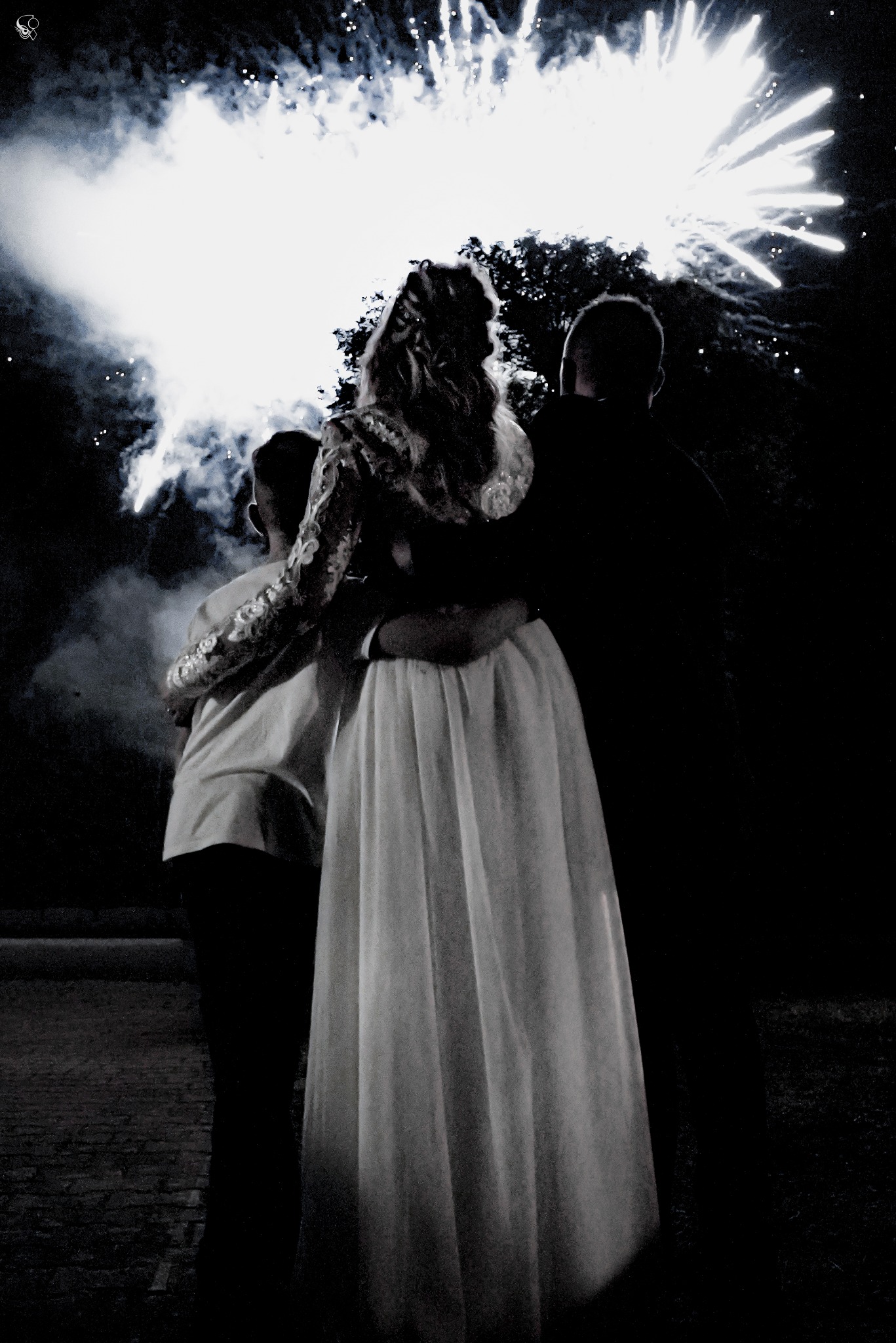 Para młoda obejmująca się, podziwiająca fajerwerki na ciemnym niebie. Panna młoda w sukni z koronkowymi rękawami, widok z tyłu, nocna sceneria.