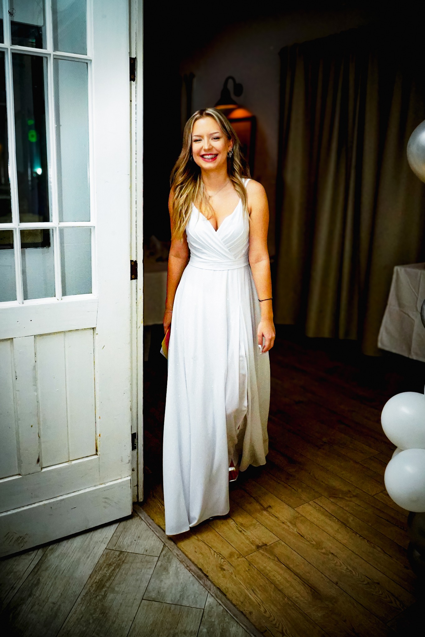 Uśmiechnięta blondynka w białej sukni wchodzi przez białe drzwi, w tle widoczne dekoracje i zasłony. Zdjęcie zrobione wewnątrz pomieszczenia.