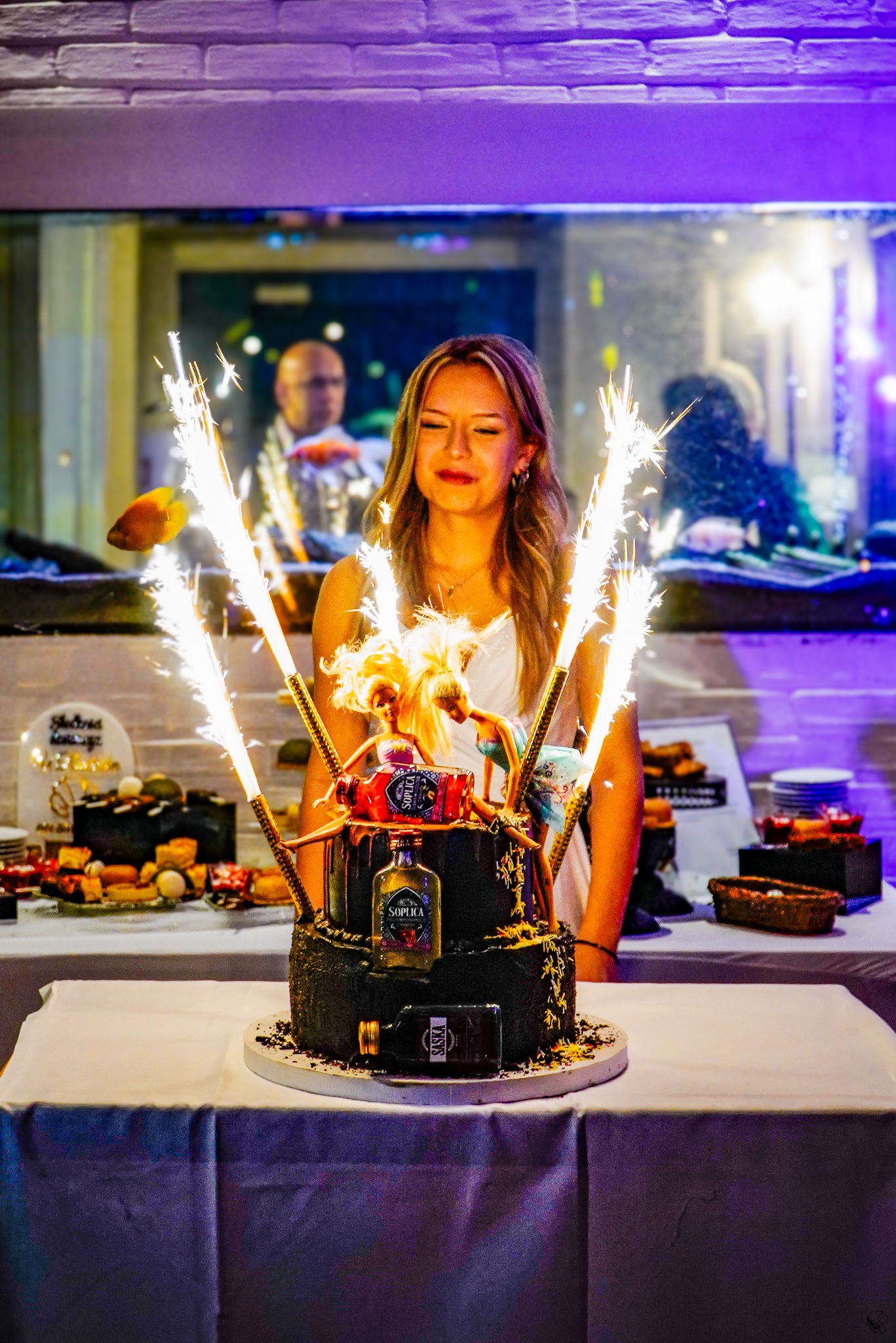 Uśmiechnięta blondynka w Koszalinie pozuje za tortem z zimnymi ogniami i dekoracjami z lalek, na tle sali z akwarium. Nietypowa dekoracja tortu.