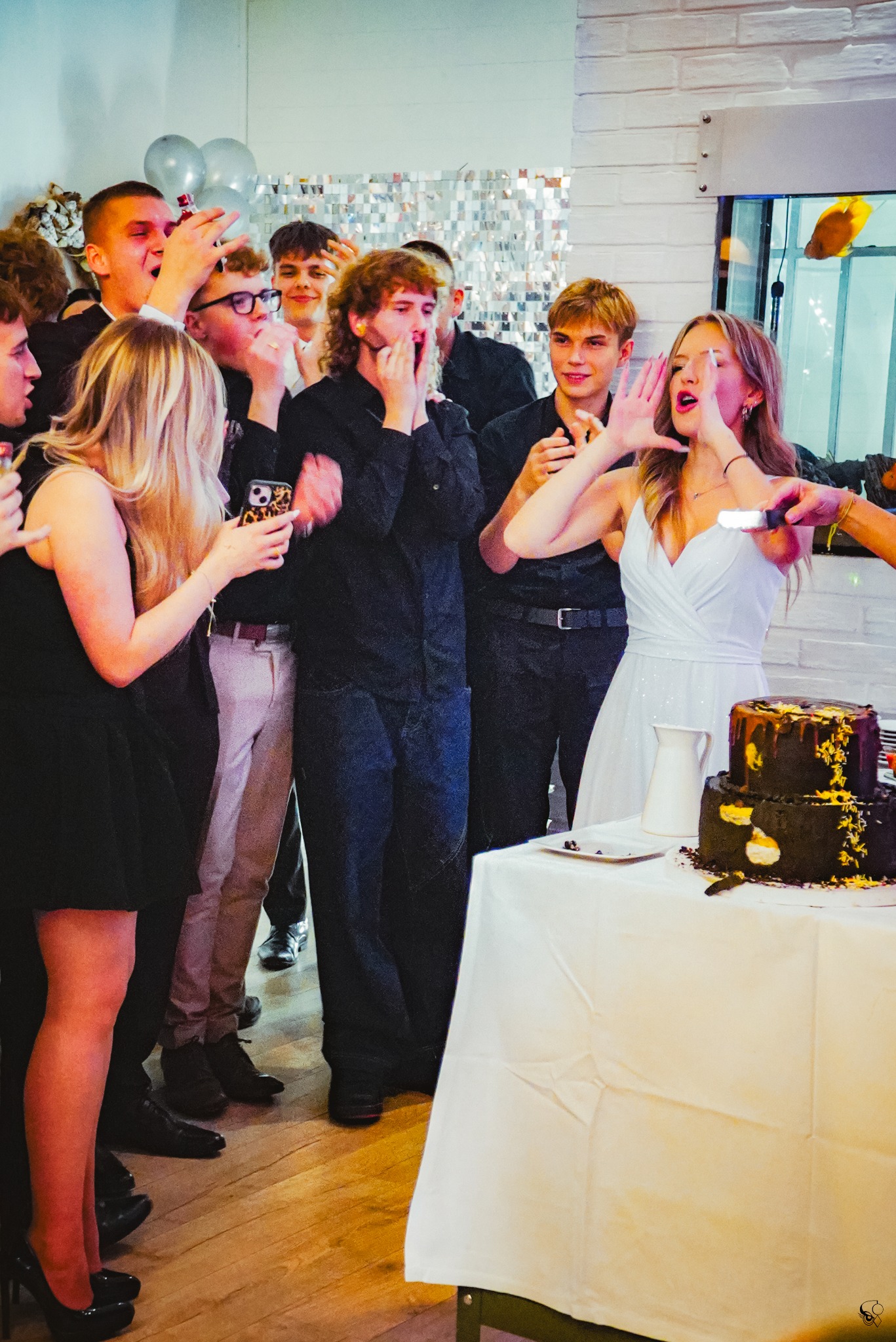 Panna młoda kroi tort weselny, otoczona gośćmi wyrażającymi radość. W tle srebrna ściana i balony. Ujęcie z bliska, dynamiczne.