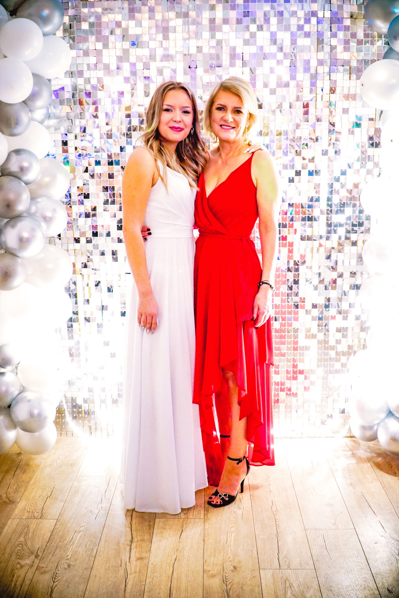 Dwie kobiety w sukniach (białej i czerwonej) pozują na tle błyszczącej ściany z balonami. Ujęcie z dołu, podkreślające sylwetki i dekoracje.