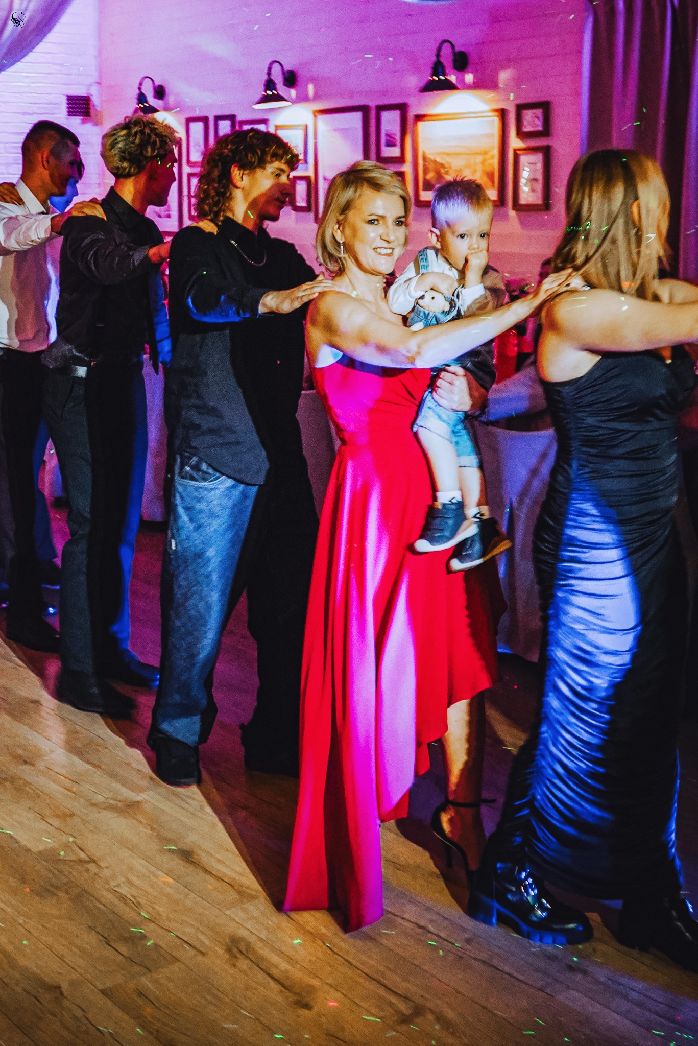 Uczestnicy wesela w trakcie tańca liniowego, kobieta w czerwonej sukni trzyma dziecko. W tle dekoracje sali i oświetlenie imprezowe.