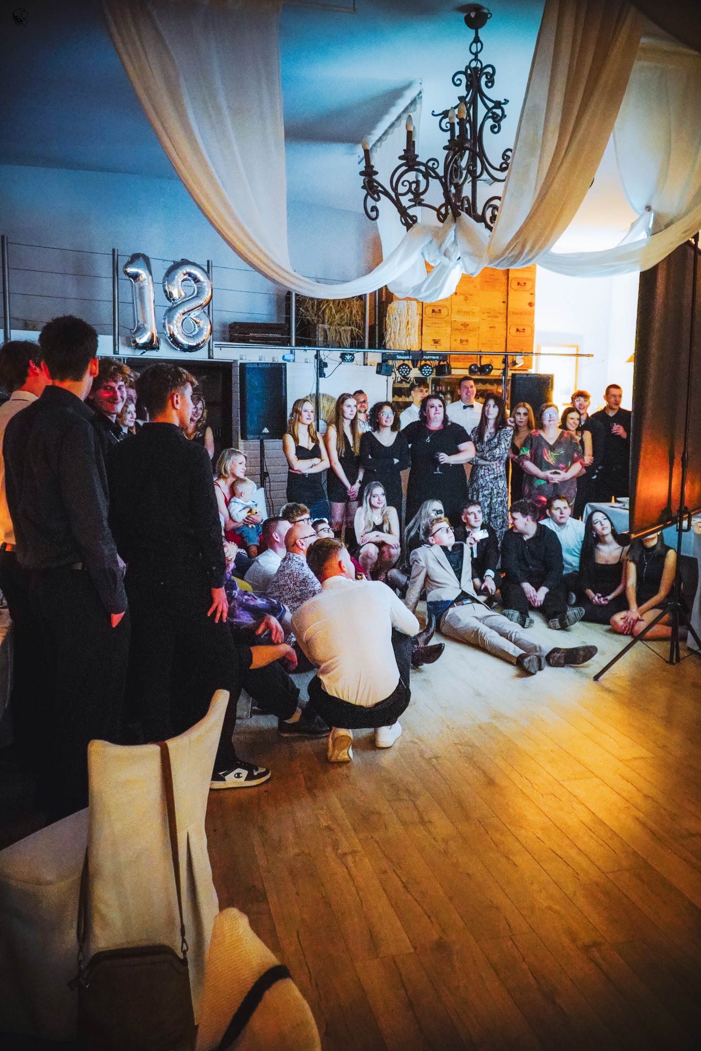 Uroczystość osiemnastych urodzin: grupa osób w różnym wieku, część siedzi na podłodze, w tle dekoracje, balony w kształcie cyfr '18', żyrandol i białe draperie.