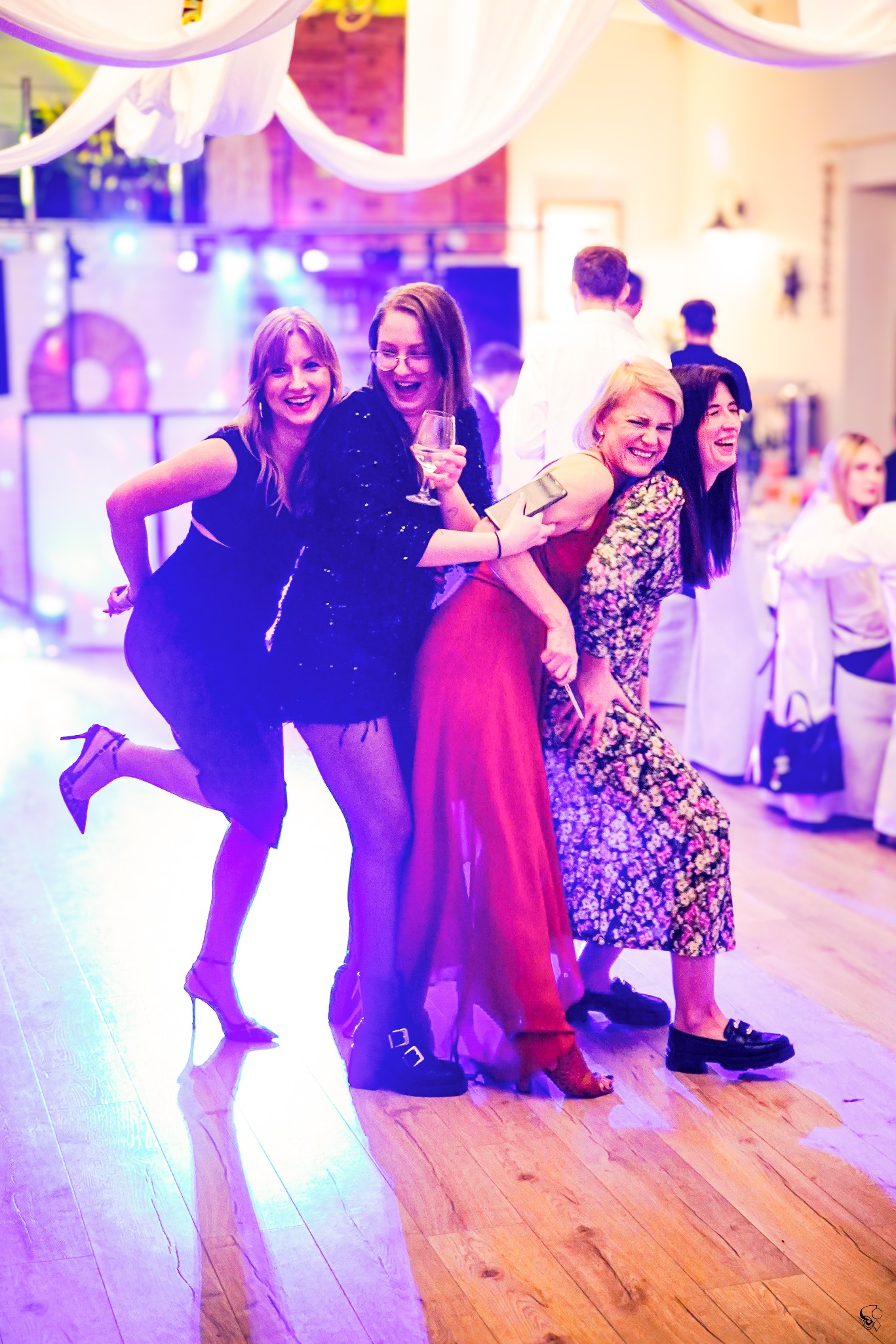 Rozbawione kobiety w różnym wieku, ubrane w eleganckie stroje, tańczą na parkiecie podczas imprezy. Widoczne światła sceniczne i parkiet z odbiciami.