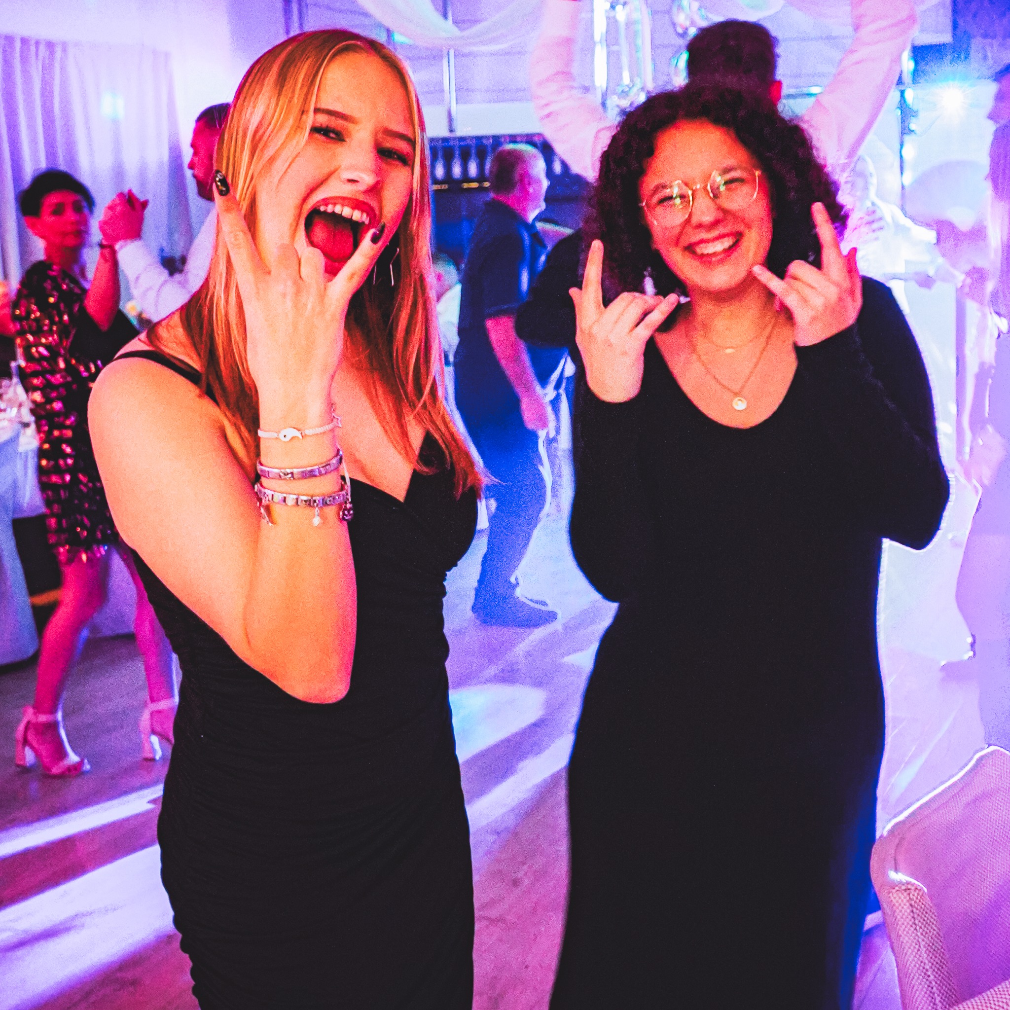 Dwie uśmiechnięte kobiety w czarnych sukienkach, pokazujące gest rockowy na imprezie z dekoracjami i tańczącymi osobami w tle, oświetlone fioletowym światłem.