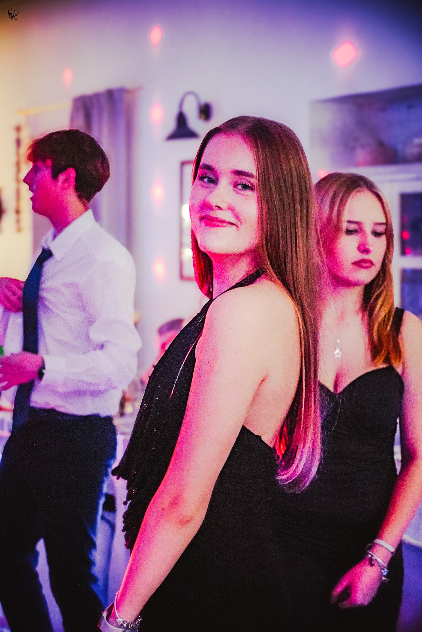 Portret uśmiechniętej młodej kobiety w czarnej sukience na tle sali balowej z dyskotekowym oświetleniem i innymi osobami w tle.