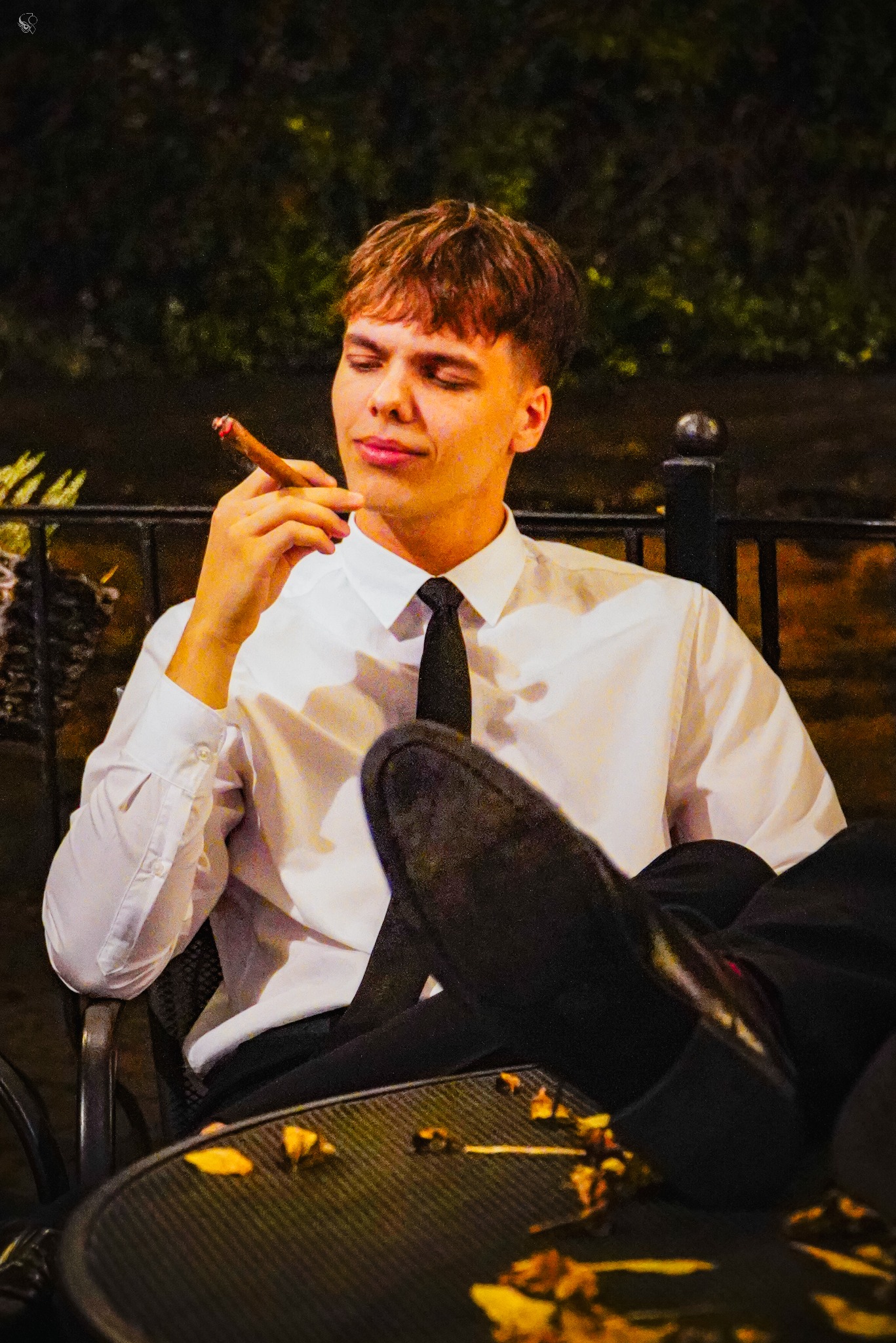 Młody mężczyzna w białej koszuli i krawacie, z nogami na stole, pali cygaro na zewnątrz. Nocne oświetlenie, metalowe krzesło i stolik z liśćmi.