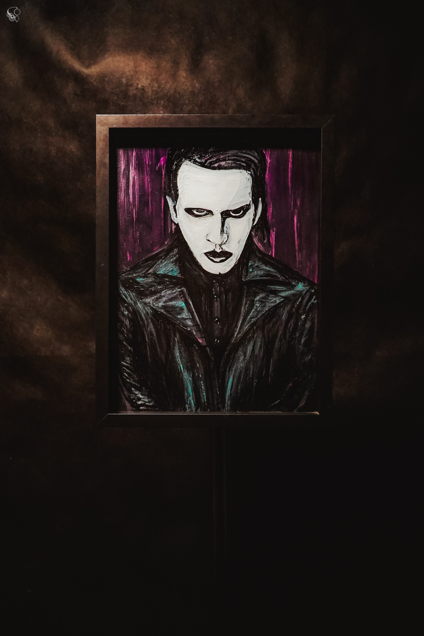 Portret w ramie, stylizowany na Marilyn Mansona, z białym makijażem i czarną skórzaną kurtką, na ciemnym tle. Wyraziste, ciemne oczy i usta.