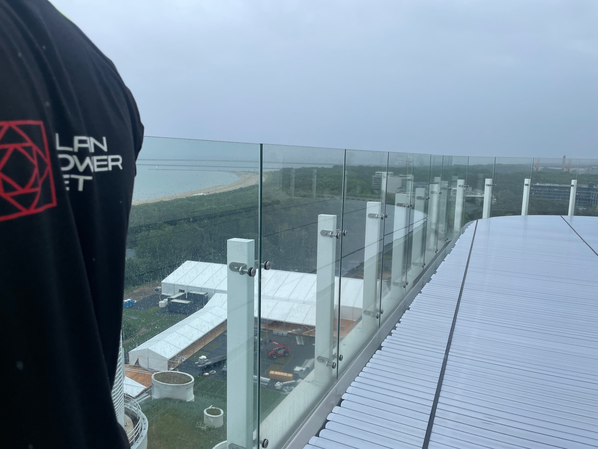Szklana balustrada na dachu budynku w deszczowy dzień, widok na morze i miasto w tle, z fragmentem osoby z logotypem firmy.
