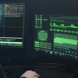 BIURO DETEKTYWISTYCZNE SIM MARIUSZ ŚCIĄŻKO - Osoba w kapturze siedzi przed dwoma monitorami wyświetlającymi zielony kod i dane. Widoczna klawiatura i mysz. Scena sugeruje cyberbezpieczeństwo lub analizę danych.