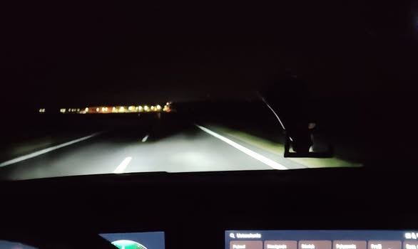 Nocna jazda samochodem z widokiem na oświetloną drogę, nagranie z wnętrza pojazdu. Widoczne światła mijanych budynków w oddali.