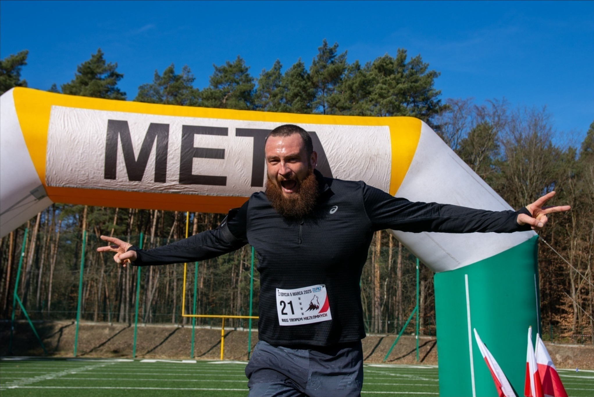 Uśmiechnięty mężczyzna z brodą przekracza linię mety biegu, w tle las i dmuchana brama z napisem META i flaga Polski. Radosny moment zwycięstwa.