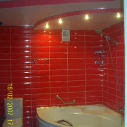 Wykończona łazienka z czerwonymi, błyszczącymi płytkami, narożną wanną z chromowanymi uchwytami i toaletą. Na suficie podwieszany sufit z oświetleniem punktowym.