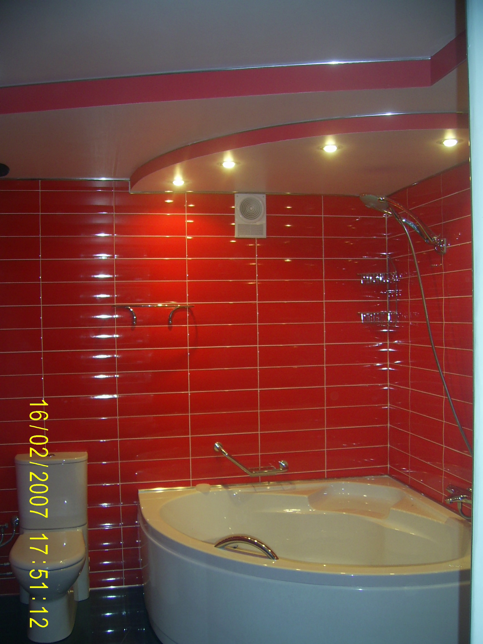 Wykończona łazienka z czerwonymi, błyszczącymi płytkami, narożną wanną z chromowanymi uchwytami i toaletą. Na suficie podwieszany sufit z oświetleniem punktowym.