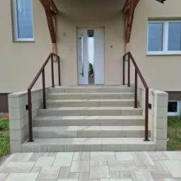 Zewnętrzne schody z jasnych płyt prowadzące do drzwi wejściowych, zabezpieczone brązowymi, minimalistycznymi barierkami po obu stronach. Widok z dołu na elewację budynku.