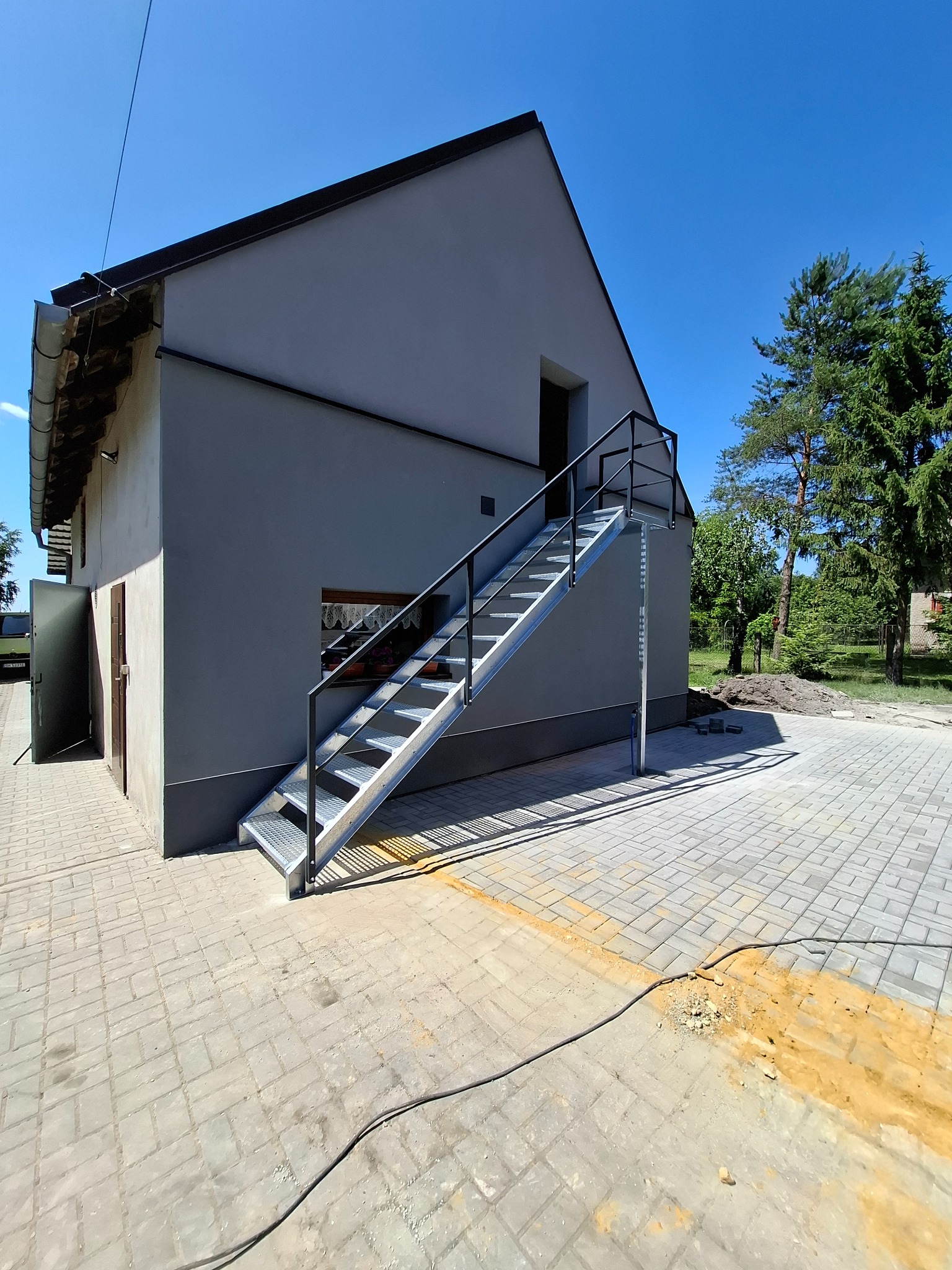 Zewnętrzne, metalowe schody z czarną poręczą prowadzące do szarego budynku z widoczną kostką brukową i zielenią w tle, w słoneczny dzień.