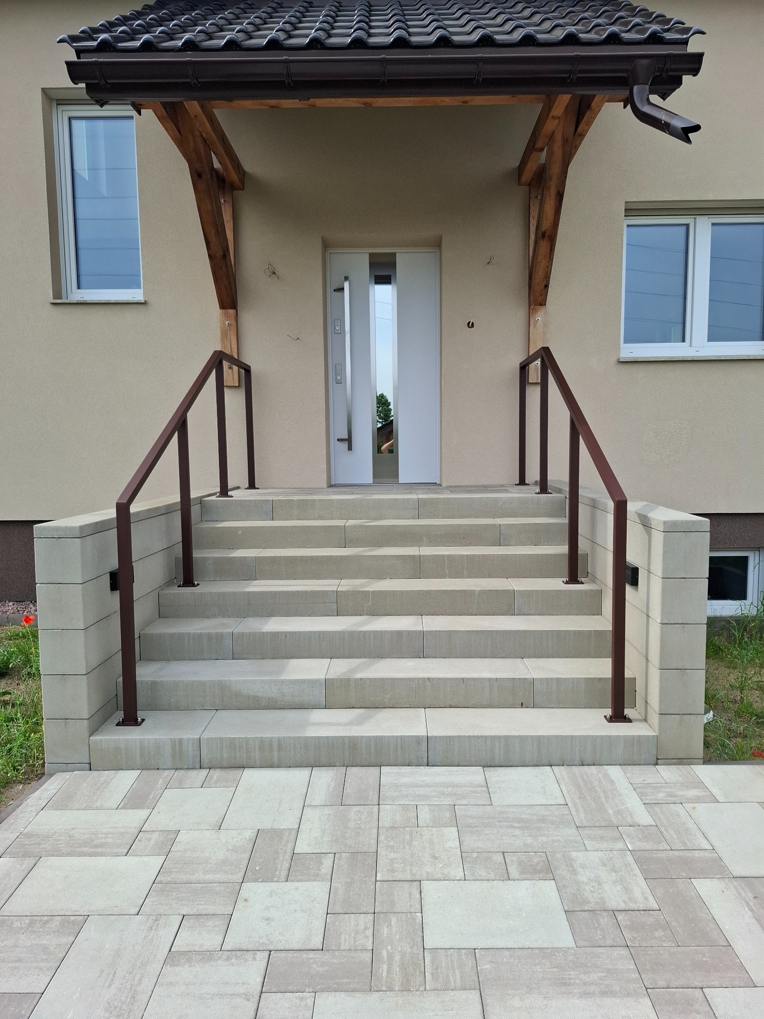 Zewnętrzne schody z jasnych płyt prowadzące do drzwi wejściowych, zabezpieczone brązowymi, minimalistycznymi barierkami po obu stronach. Widok z dołu na elewację budynku.