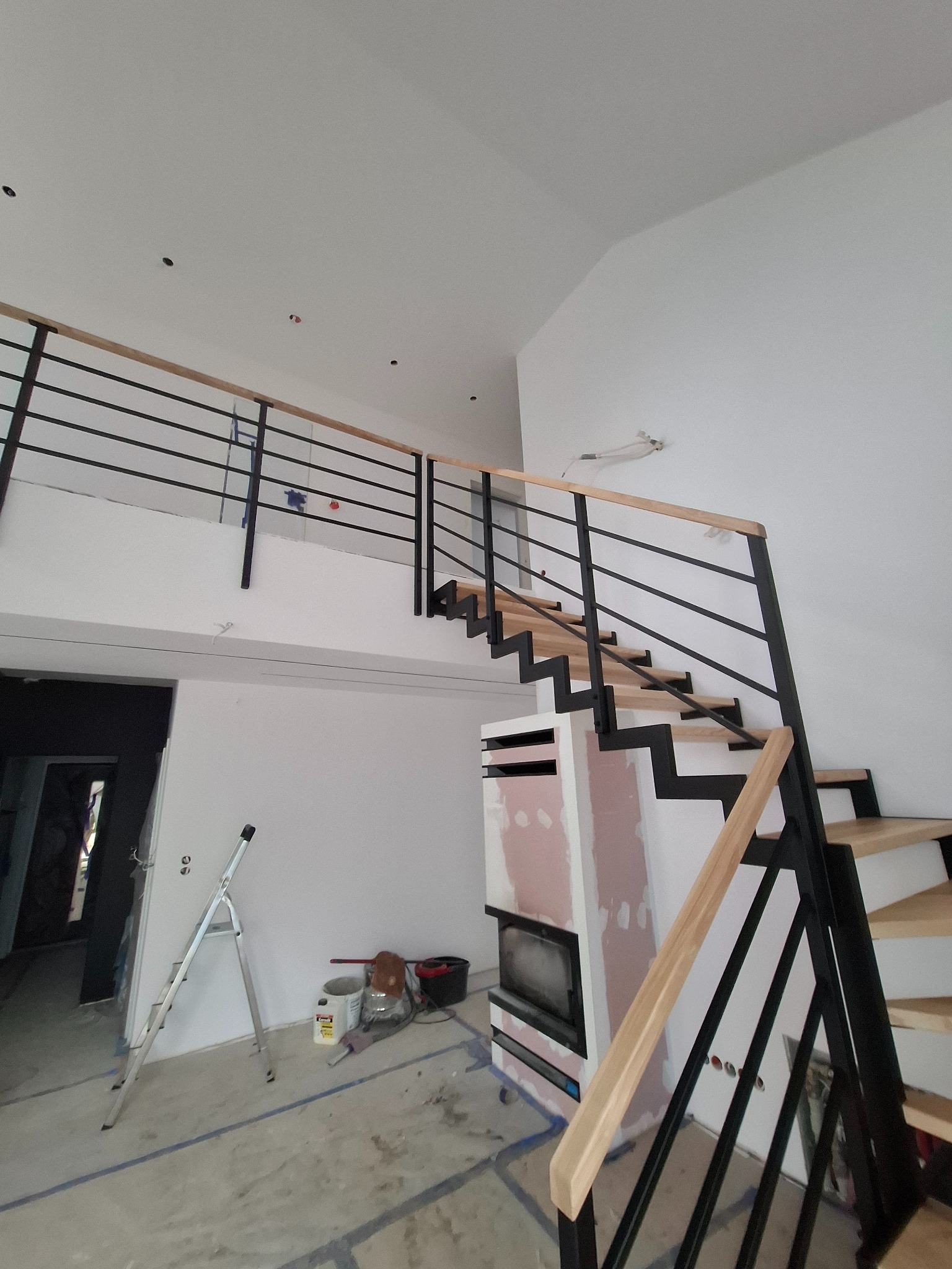 Nowoczesne schody z czarną metalową konstrukcją i drewnianymi stopniami w jasnym wnętrzu z kominkiem. Widok z dołu, podkreślający geometrię i minimalistyczny design.