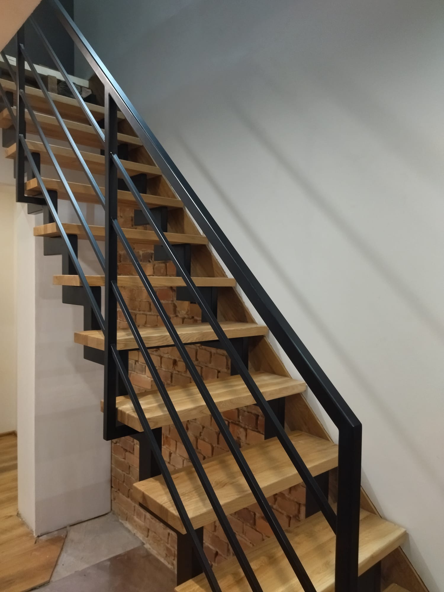Nowoczesne schody z drewnianymi stopniami i czarną metalową konstrukcją na tle ceglanej ściany, widok z dołu, oświetlenie dzienne.