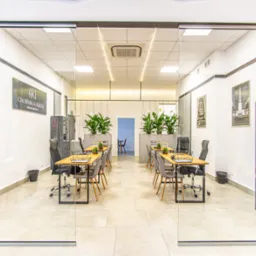 Nowoczesne biuro agencji nieruchomości z przeszklonymi ścianami, drewnianymi biurkami i roślinami doniczkowymi. Widoczne plakaty na ścianach i minimalistyczny design.