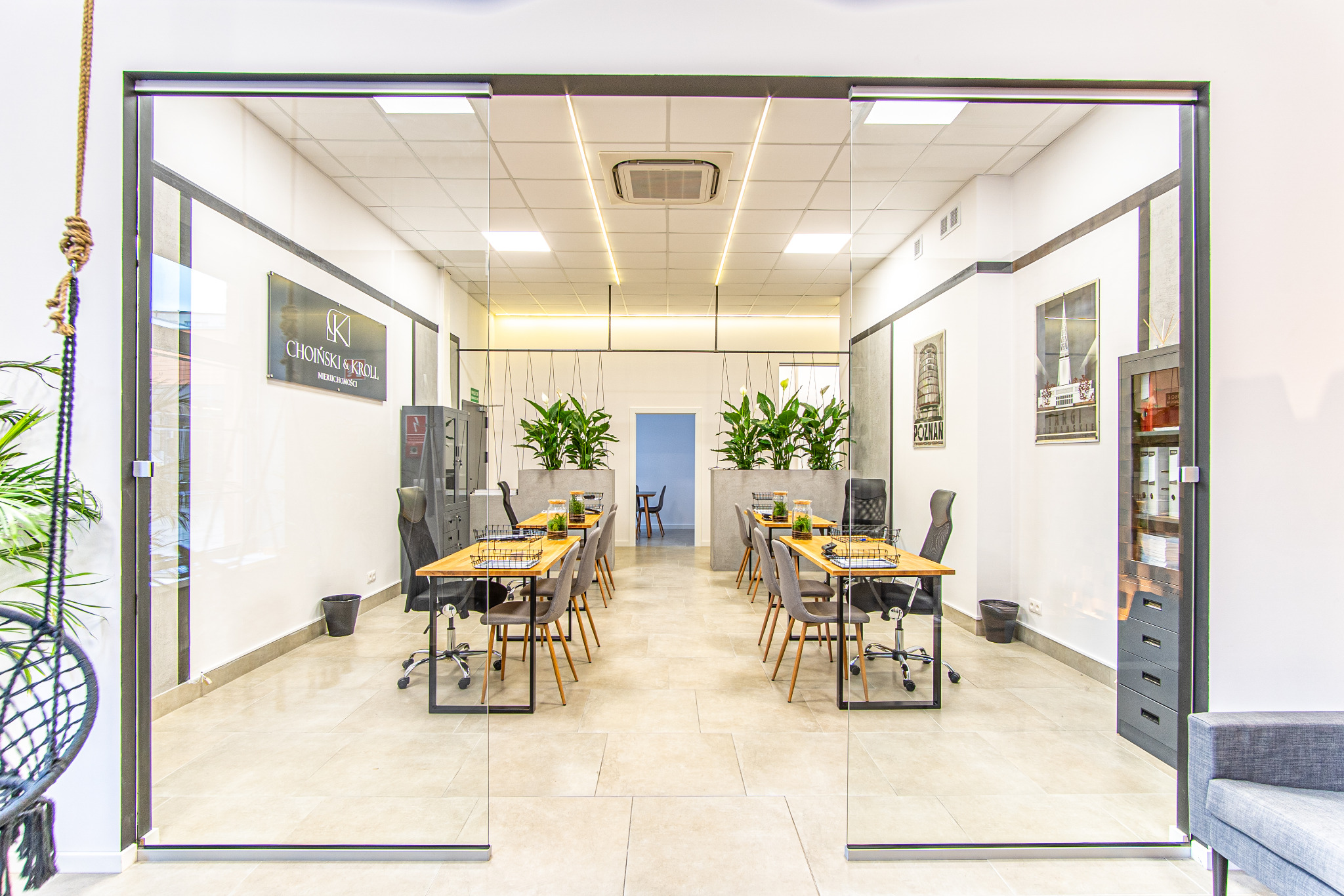 Nowoczesne biuro agencji nieruchomości z przeszklonymi ścianami, drewnianymi biurkami i roślinami doniczkowymi. Widoczne plakaty na ścianach i minimalistyczny design.