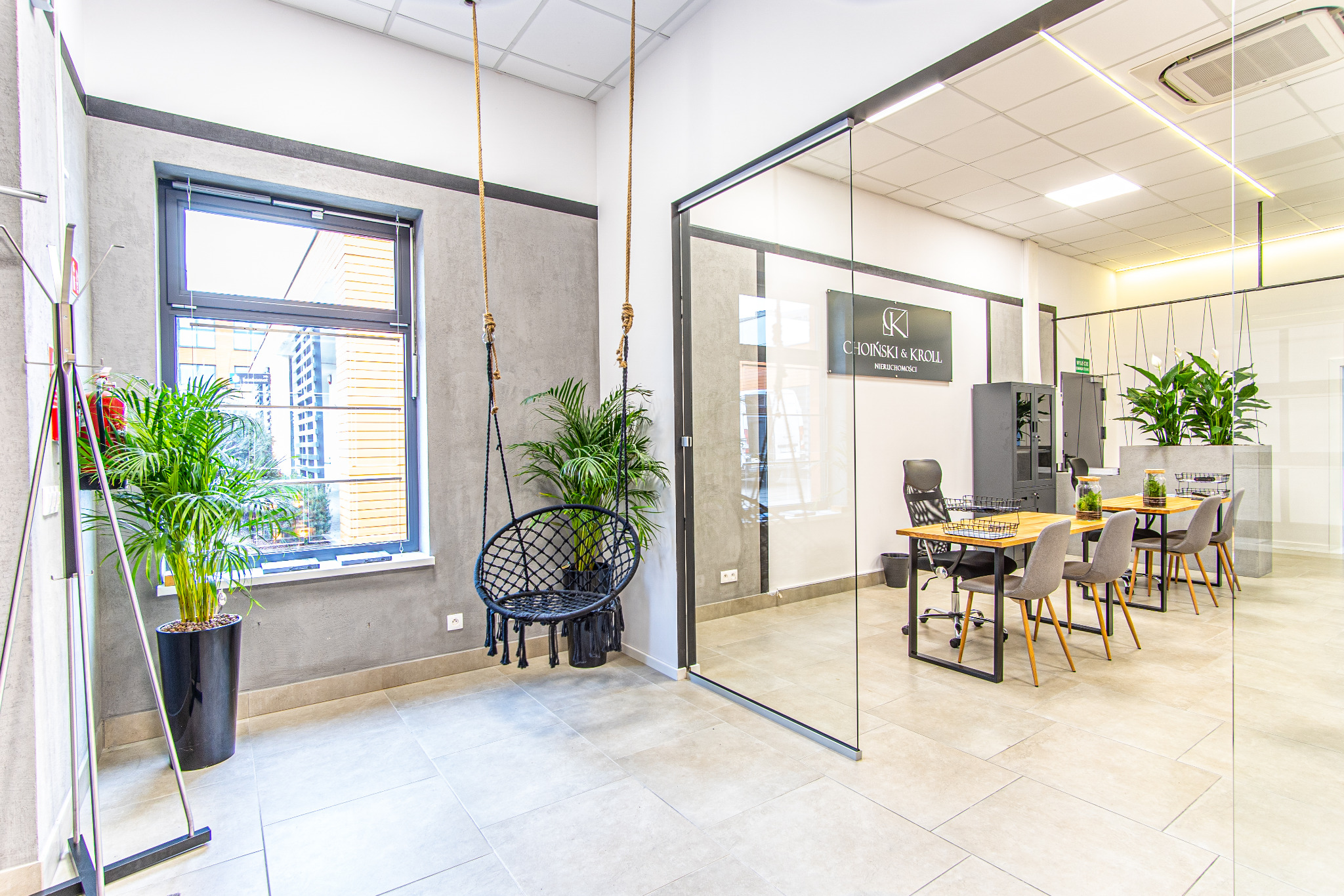 Nowoczesne biuro agencji nieruchomości z wiszącym fotelem, roślinami i przeszkloną salą konferencyjną. Widoczne logo firmy Choinski & Kroll Nieruchomości.